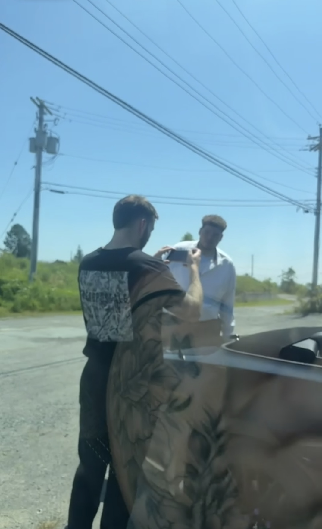 Crew member filming an actor beside a convertible on a sunny roadside